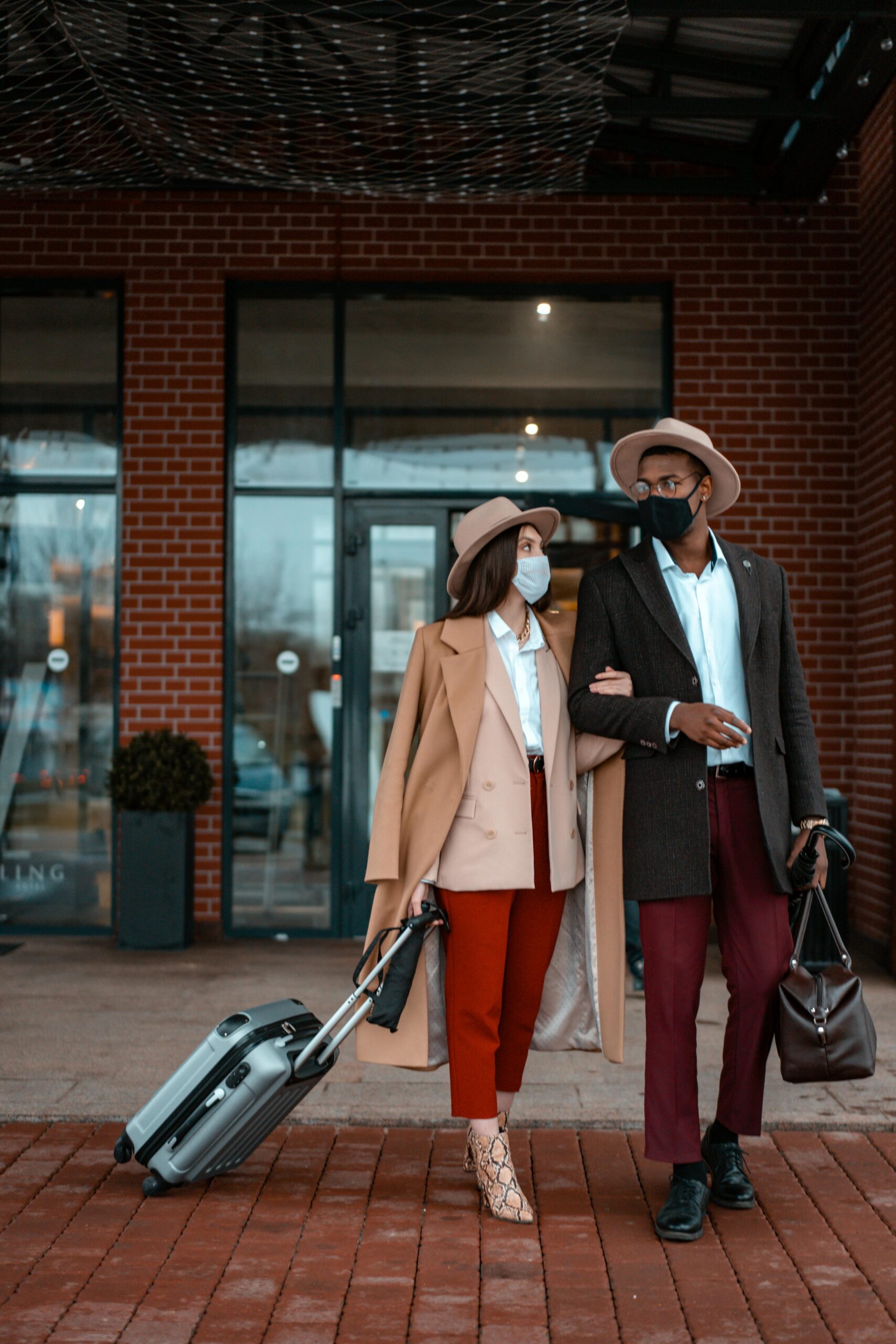 Fashionable couple with face masks traveling with luggage outside a hotel.